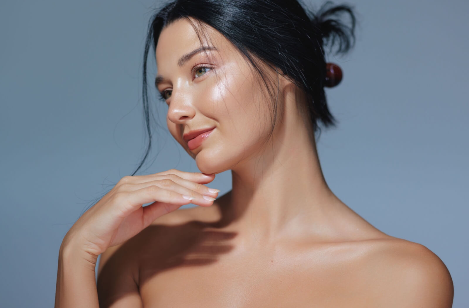 A person with glowing skin posing gracefully with the hand under her chin, looking to the side against a soft blue background.
